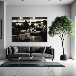 May include: A large framed print of a restaurant scene hangs above a gray sofa. Two men sit at a table, with a kitchen and staff visible in the background. A potted tree stands to the right of the sofa.