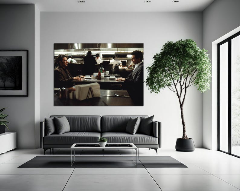 May include: A large framed print of a restaurant scene hangs above a gray sofa. Two men sit at a table, with a kitchen and staff visible in the background. A potted tree stands to the right of the sofa.