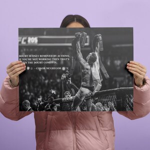 May include: Black and white poster featuring a victorious athlete holding championship belts. The poster includes the text "Doubt is only removed by actions. If you're not working then that's when the doubt comes in." by Conor McGregor.