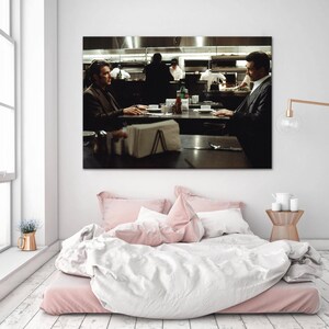 May include: A framed print of a scene from a movie, depicting two men seated at a table in a restaurant. The print is mounted above a bed with pink bedding and a white duvet. A gold lamp and small side table are also visible.