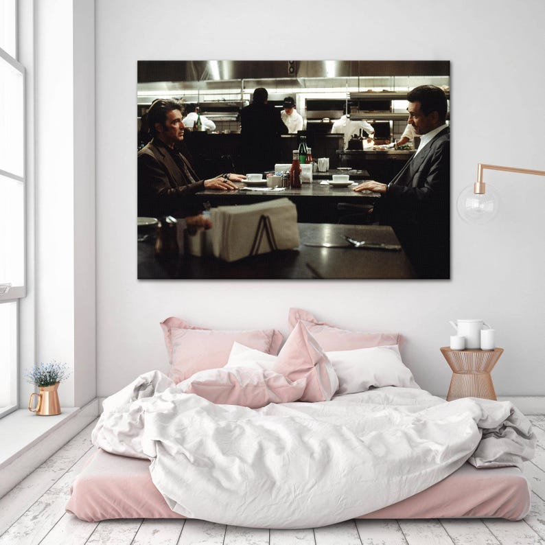 May include: A framed print of a scene from a movie, depicting two men seated at a table in a restaurant. The print is mounted above a bed with pink bedding and a white duvet. A gold lamp and small side table are also visible.