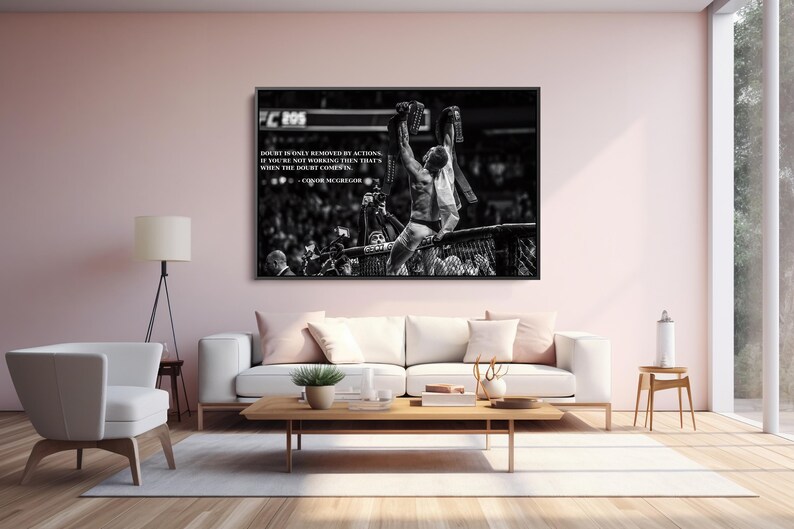 May include: Black and white framed print of a mixed martial arts fighter holding championship belts above his head. The print includes a quote by Conor McGregor. The image is displayed above a white sofa.