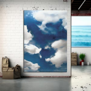 May include: A large canvas print showcasing a blue sky with white, fluffy clouds. The artwork is mounted on a white brick wall. A view of the sea is visible through a window in the background.