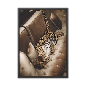 May include: A framed art print showing a leopard reclining on a tufted, brown velvet sofa. The leopard's coat is patterned with spots, and it is positioned with its head towards the viewer. The sofa has several cushions and a black chain.