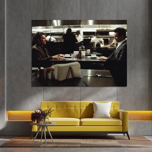 May include: A large framed print of a scene from a movie, depicting two men seated at a table in a restaurant. The print hangs above a yellow sofa with a white pillow. A small side table with flowers sits in front of the sofa.