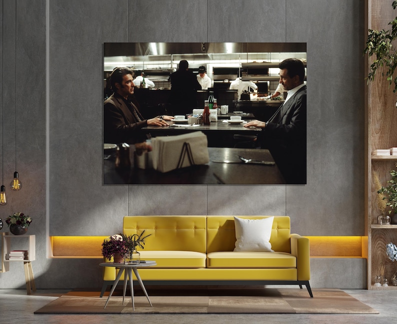 May include: A large framed print of a scene from a movie, depicting two men seated at a table in a restaurant. The print hangs above a yellow sofa with a white pillow. A small side table with flowers sits in front of the sofa.