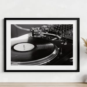May include: Black and white framed photograph of a turntable with a vinyl record. The turntable is in focus, with the record's grooves and center label visible. The background shows a mixing console, suggesting a DJ setup.
