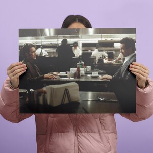 May include: A framed print depicting a scene from a film, featuring two men seated at a table in a restaurant. The image is in muted tones, with a focus on the characters and the setting. Napkins, condiments, and tableware are visible on the table.
