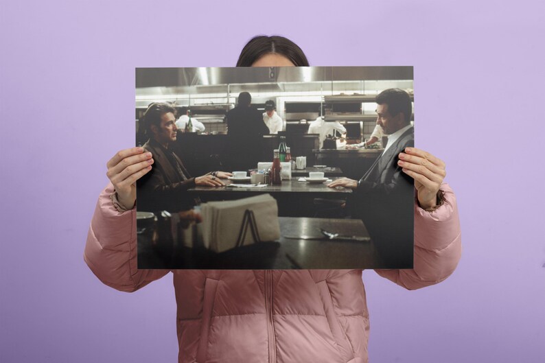 May include: A framed print depicting a scene from a film, featuring two men seated at a table in a restaurant. The image is in muted tones, with a focus on the characters and the setting. Napkins, condiments, and tableware are visible on the table.