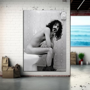 Puede incluir: Fotografía en blanco y negro de una persona sentada en un inodoro. La persona tiene el pelo largo y rizado y un bigote. Un teléfono antiguo está montado en la pared detrás de ellos.