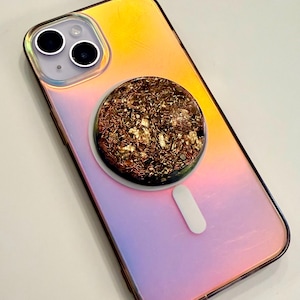 May include: Iridescent phone case with a circular, decorative element. The case transitions from yellow to pink and purple. The circular element is filled with copper-colored wire and gold flakes, attached to a white, rectangular grip.