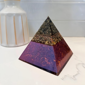 May include: A handcrafted pyramid sculpture with a layered design. The top section is dark with gold accents, transitioning to a deep purple base. The pyramid is displayed on a white surface.