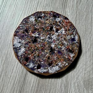 May include: Round, handcrafted resin coaster with a mix of silver foil, copper wire, and small gemstones. The coaster has a light brown edge and a dark purple and silver center. A unique, decorative item for home decor.