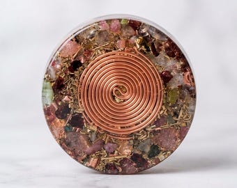 May include: A circular resin disc featuring a central copper wire spiral coil surrounded by embedded fragments of pink, red, and clear gemstones along with small metallic shavings, creating a textured, multi-colored appearance.