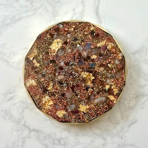May include: A round, decorative object with a gold-colored rim. The interior is filled with a mix of small stones, copper wire, and gold leaf. The stones are various colors, including red, purple, and white. The object is set against a white marble background.