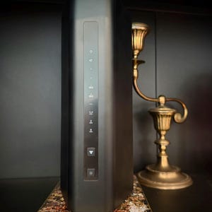 May include: A black rectangular electronic device with indicator lights and buttons, resting on a decorative stand with gold trim and a patterned surface. A gold candlestick holder is to the right.