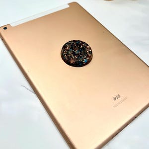 May include: A rose gold tablet with a circular, dark-colored phone grip attached to the back. The grip features a textured, multi-colored inlay with flecks of copper, blue, and black embedded within a glossy, dark resin surface.
