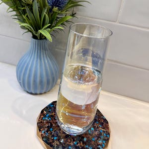May include: A clear glass filled with water rests on a decorative coaster with a gold rim and a mosaic of brown and blue stones. A blue vase with blue thistle flowers is in the background. The coaster is approximately 4 inches in diameter.