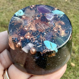 May include: A round, dark-colored resin art piece with embedded gemstones and copper wire. The top surface features amethyst, turquoise, and clear stones, along with copper beads and black beads. The piece is held in a hand.