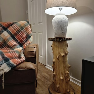 Puede incluir: Lámpara de pie rústica con base de cerámica texturizada blanquecina y pantalla beige. La lámpara se apoya en un soporte de madera hecho de un tronco de árbol, de unos 122 cm de altura, aportando un toque natural.