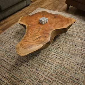 May include: A distinctive, live-edge wooden coffee table with an irregular shape. The table is a warm, medium brown colour, displaying the wood grain. A small stack of square coasters sits on the table's surface. The table rests on a patterned rug.