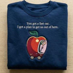 Könnte beinhalten: Ein marineblaues Sweatshirt mit Rundhalsausschnitt und weißem Sticktext: "You got a fast car. I got a plan to get us out of here." Unter dem Text befindet sich ein gestickter roter Apfel in Form eines Autos mit einem Wurm als Fahrer.