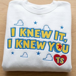 May include: A white crewneck sweatshirt featuring embroidered text that reads I KNEW IT, I KNEW YOU in yellow with blue outlines. The design includes small blue stars, white cloud outlines, a colorful ball with a red star, and a red heart with TS.