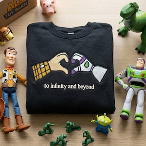 Puede incluir: Una sudadera negra de cuello redondo con un dise&ntilde;o bordado de dos manos formando un coraz&oacute;n, una con estampado de camisa de vaquero y la otra de traje de guardi&aacute;n espacial, con el texto to infinity and beyond bordado debajo.