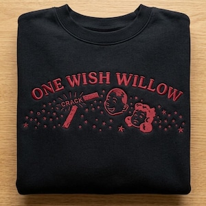 May include: A black crewneck sweatshirt featuring red embroidery. The text reads ONE WISH WILLOW with the word CRACK next to a broken stick. The design includes two cartoon faces and small star shapes scattered across the chest area.