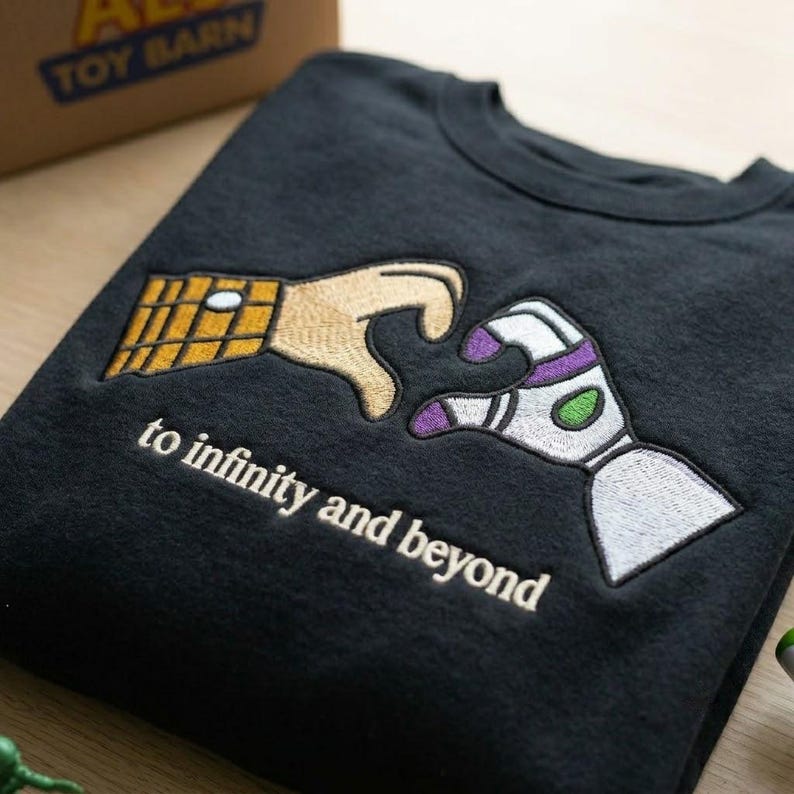 Puede incluir: Una sudadera negra de cuello redondo con un dise&ntilde;o bordado de dos manos que se acercan. Una mano tiene un patr&oacute;n de cuadros amarillos y la otra es blanca, morada y verde. El texto "to infinity and beyond" est&aacute; bordado en la parte inferior.
