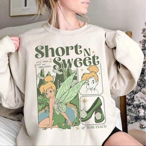 May include: Beige sweatshirt featuring a graphic of Tinkerbell with the text "Short N Sweet". The design includes illustrations of Tinkerbell, a landscape, and green high heels. The phrase "I leave quite an impression" is also included.