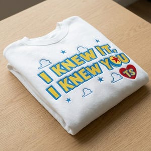May include: A white crewneck sweatshirt featuring embroidered text that reads I KNEW IT, I KNEW YOU TS in yellow with blue outlines. The design includes small blue stars, white cloud outlines, and a red heart containing a yellow star icon.