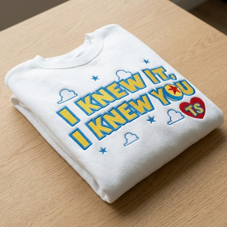 May include: A white crewneck sweatshirt featuring embroidered text that reads I KNEW IT, I KNEW YOU TS in yellow with blue outlines. The design includes small blue stars, white cloud outlines, and a red heart containing a yellow star icon.
