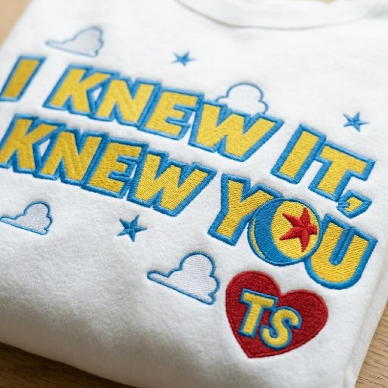 May include: A white sweatshirt featuring yellow embroidered text that reads I KNEW IT, KNEW YOU with blue outlines. The design includes a blue and yellow ball with a red star, a red heart with yellow TS letters, and small blue clouds and stars.