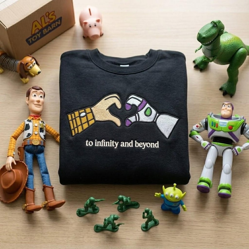 Puede incluir: Una sudadera negra de cuello redondo con un dise&ntilde;o bordado de dos manos formando un coraz&oacute;n, una con estampado de camisa de vaquero y la otra de traje de guardi&aacute;n espacial, con el texto to infinity and beyond bordado debajo.