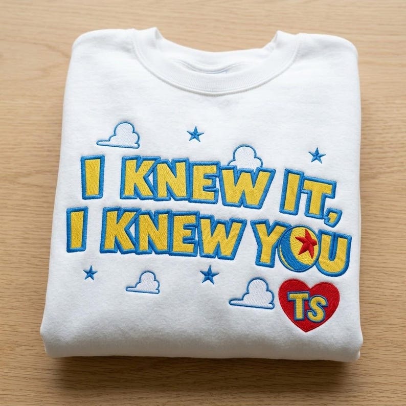 May include: A white crewneck sweatshirt featuring embroidered text that reads I KNEW IT, I KNEW YOU in yellow with blue outlines. The design includes small blue stars, white cloud outlines, a colorful ball with a red star, and a red heart with TS.