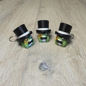 May include: Three green frog keychains wearing black top hats with white bands, bow ties, and monocles. The frog keychains are on a wooden surface.