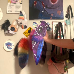 May include: A purple and blue decorative wing with white accents is held up against a wall. The wing has a geometric design and is made of paper or cardboard. Other items on the wall include artwork, a cat-themed sign, and a rainbow-colored tail.