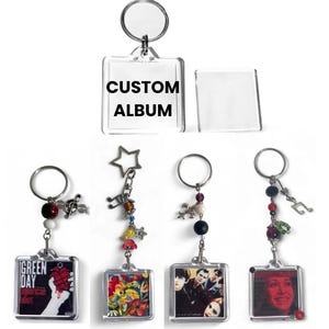 Peut inclure: Une collection de porte-clés carrés en acrylique avec des anneaux métalliques et des perles décoratives. Un porte-clés affiche le texte CUSTOM ALBUM. D'autres présentent diverses pochettes d'album, dont Green Day, avec des breloques en forme de note de musique et d'étoile.