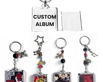 May include: A collection of square acrylic keychains featuring metal rings and decorative beads. One keychain displays the text CUSTOM ALBUM. Others feature various album cover art, including Green Day, with musical note and star charms attached.