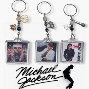 May include: Three clear acrylic keychains featuring images of Michael Jackson album covers. Each keychain has a silver ring and decorative charms, including a peace sign, a microphone, and beads. The Michael Jackson signature logo is below.