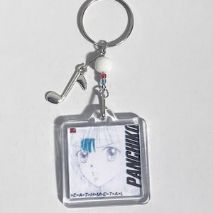 May include: A clear acrylic keychain with a square pendant featuring a manga-style illustration of a person's face. The keychain includes the text "PANCHIKO" and "E>A>T>H>M>E>T>A>L". It has a silver-coloured key ring, a musical note charm, and a white bead.