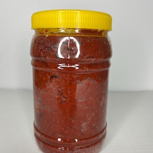 Pode incluir: Um frasco cilíndrico de plástico transparente preenchido com uma pasta espessa de cor vermelho-escura e textura granulada. O recipiente possui uma tampa de rosca amarela brilhante com estrias e uma costura vertical ao longo do corpo.