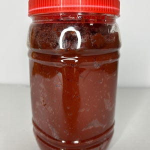 Op de afbeelding: Een doorzichtige plastic pot gevuld met een donkerrode, brokkelige substantie. De pot heeft een rode schroefdop. De inhoud lijkt op een dikke pasta of saus, mogelijk een kruiderij of voedingsproduct.
