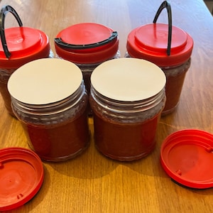 Op de afbeelding: Een verzameling glazen potten gevuld met een roodbruine substantie, waarschijnlijk voedsel. De potten hebben rode deksels, sommige met zwarte handvatten. Witte zegels bedekken de openingen van sommige potten. De potten zijn gerangschikt op een houten oppervlak.