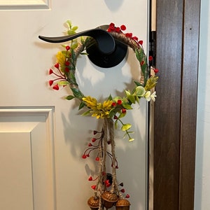 Może przedstawiać: Jesienna wieniec na drzwi z podstawą z brązowego sznurka, ozdobiony czerwonymi jagodami, zielonymi liśćmi i żółtymi kwiatami. Z dołu wieńca zwisa sznur brązowych żołędzi.