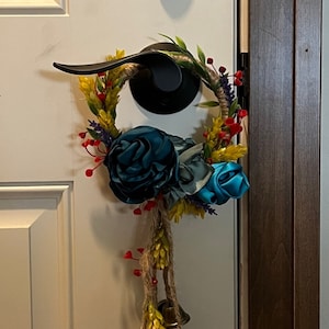 Może przedstawiać: Wianek kwiatowy w kolorze niebieskim i żółtym z sznurkiem jutowym i dzwonkami wiszącymi od dołu. Wianek jest zawieszony na białych drzwiach z czarnym klamką.