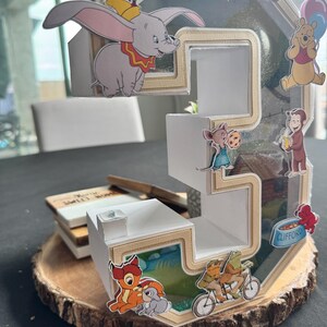 May include: A three-dimensional number three display featuring the name GRAYSON at the top. The white structure is decorated with various cartoon character cutouts including Dumbo, Winnie the Pooh, Curious George, Clifford, Bambi, and Thumper.