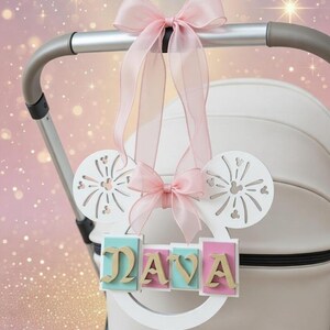 Puede incluir: Accesorio blanco para cochecito con el nombre "DAVA" en letras doradas sobre un marco blanco. Decorado con lazos rosas y recortes con forma de orejas de Mickey. Fondo rosa y dorado suave, ideal para un bebé.