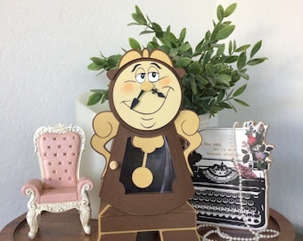 May include: A brown and tan paper craft clock figure with a smiling face, black hands, and a pendulum, positioned next to a miniature pink tufted chair and a black and white typewriter illustration with the text the end, we'll all become stories.